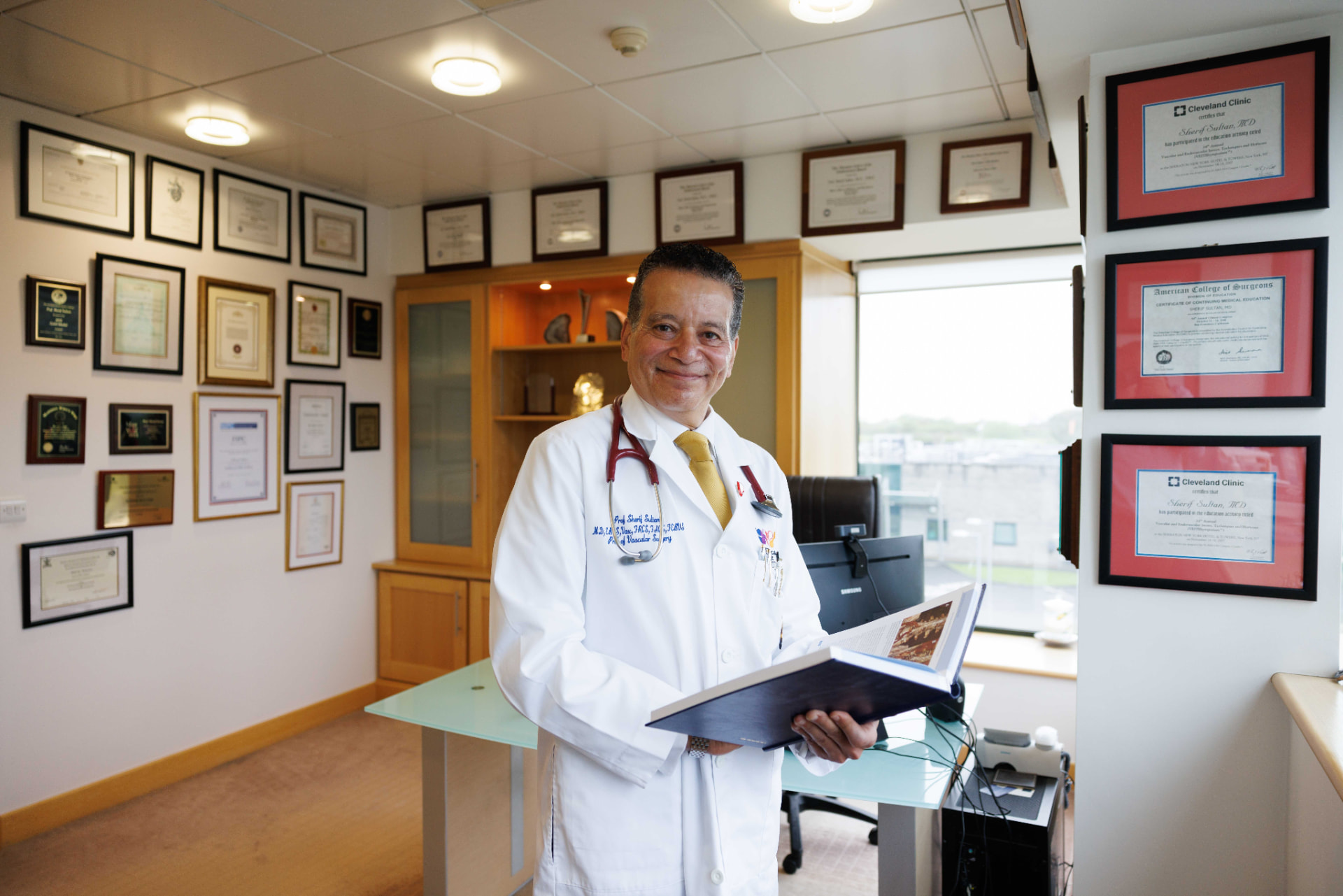 Professor Sherif Sultan in his white coat, standing in his Galway Clinic office surrounded by qualifications and awards